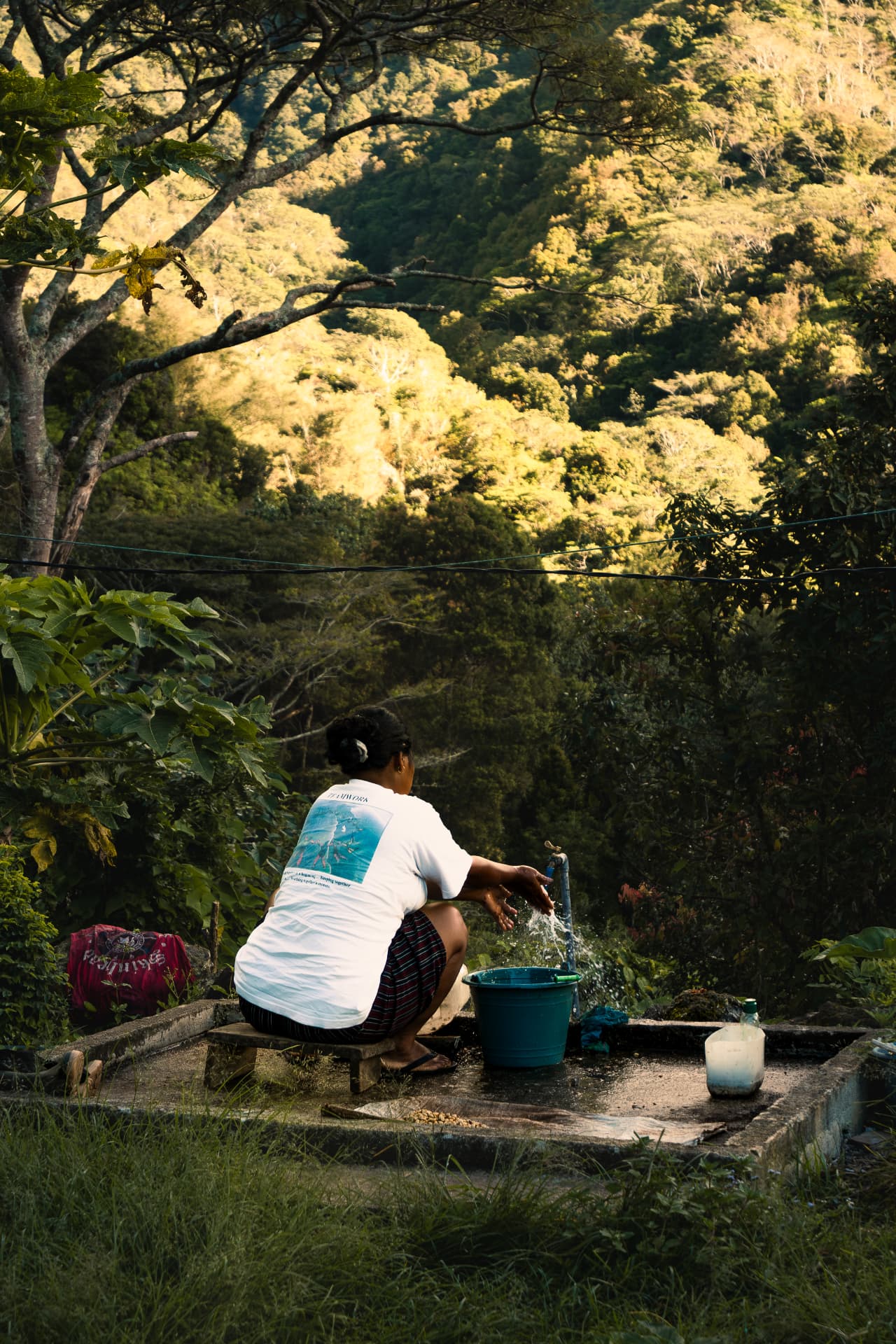 Woman washing beside a bucket at an outdoor tap with forested hills behind