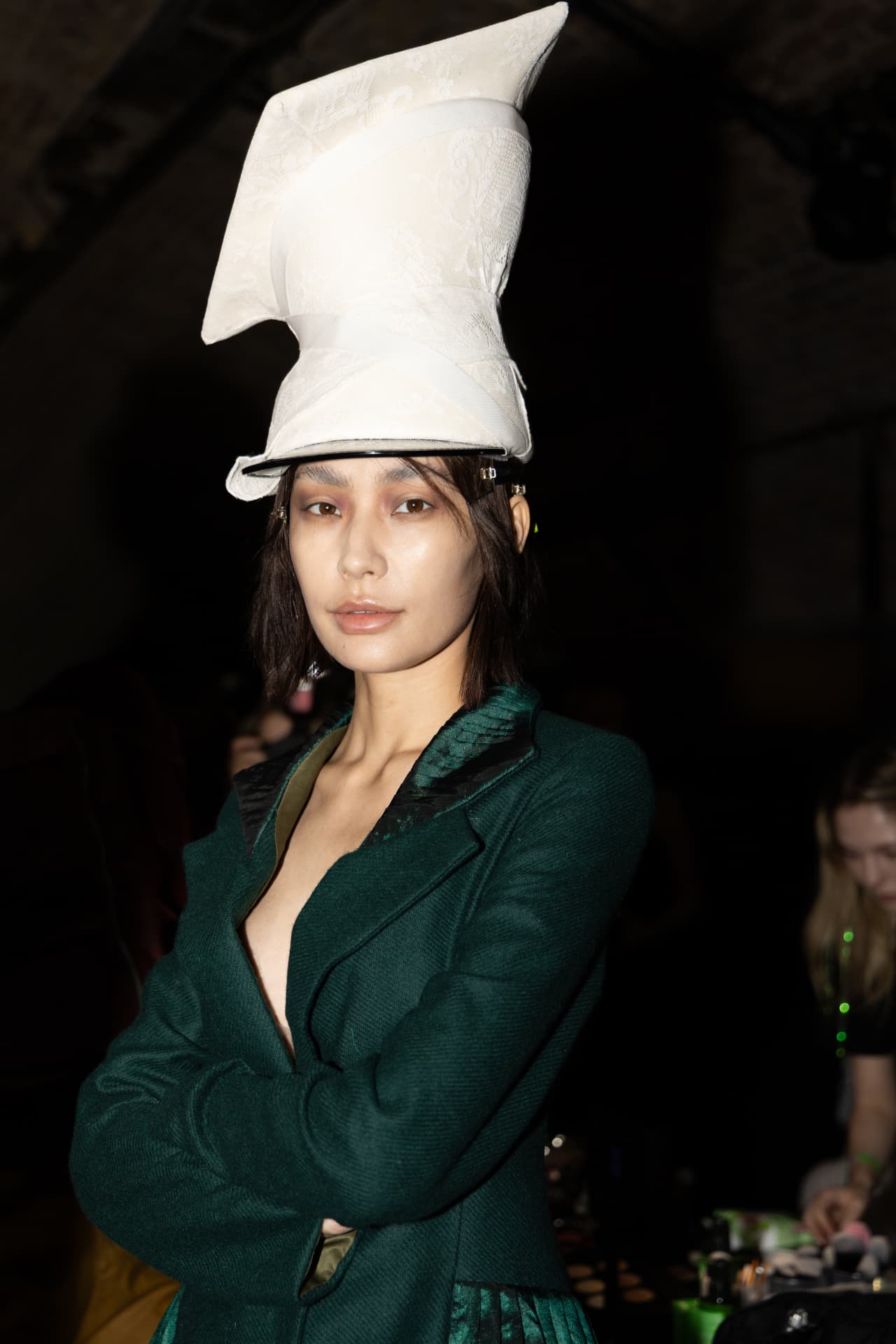 Model in a sculptural white headpiece and emerald jacket backstage
