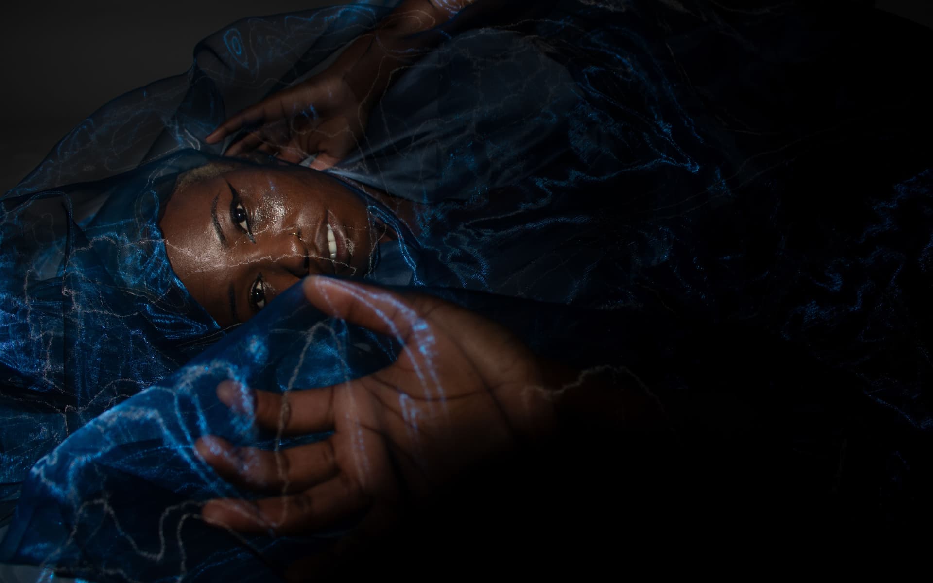 Model lying beneath blue organza reaching toward the camera