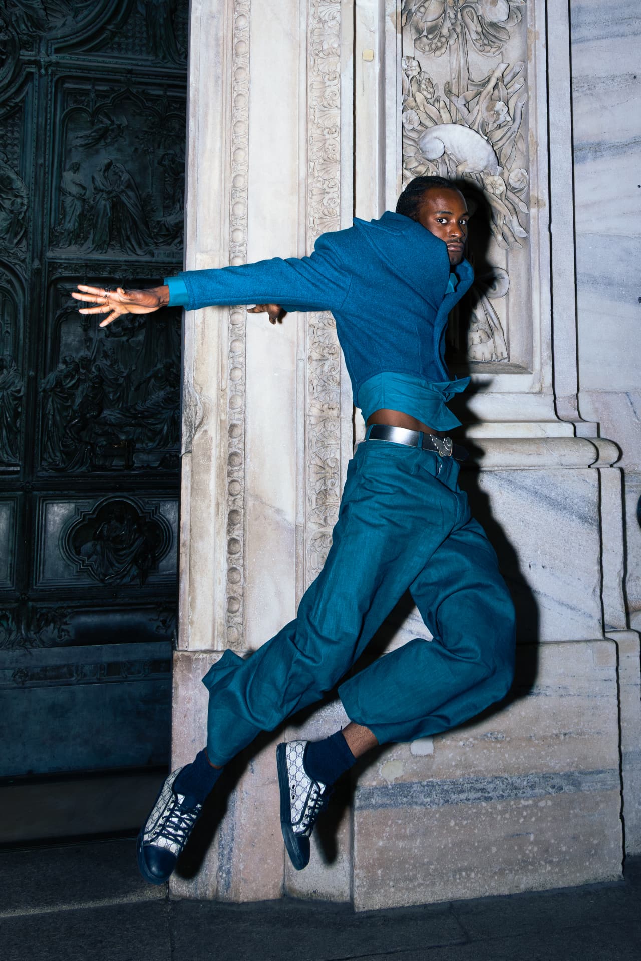 Model in a teal suit leaping against the stone facade