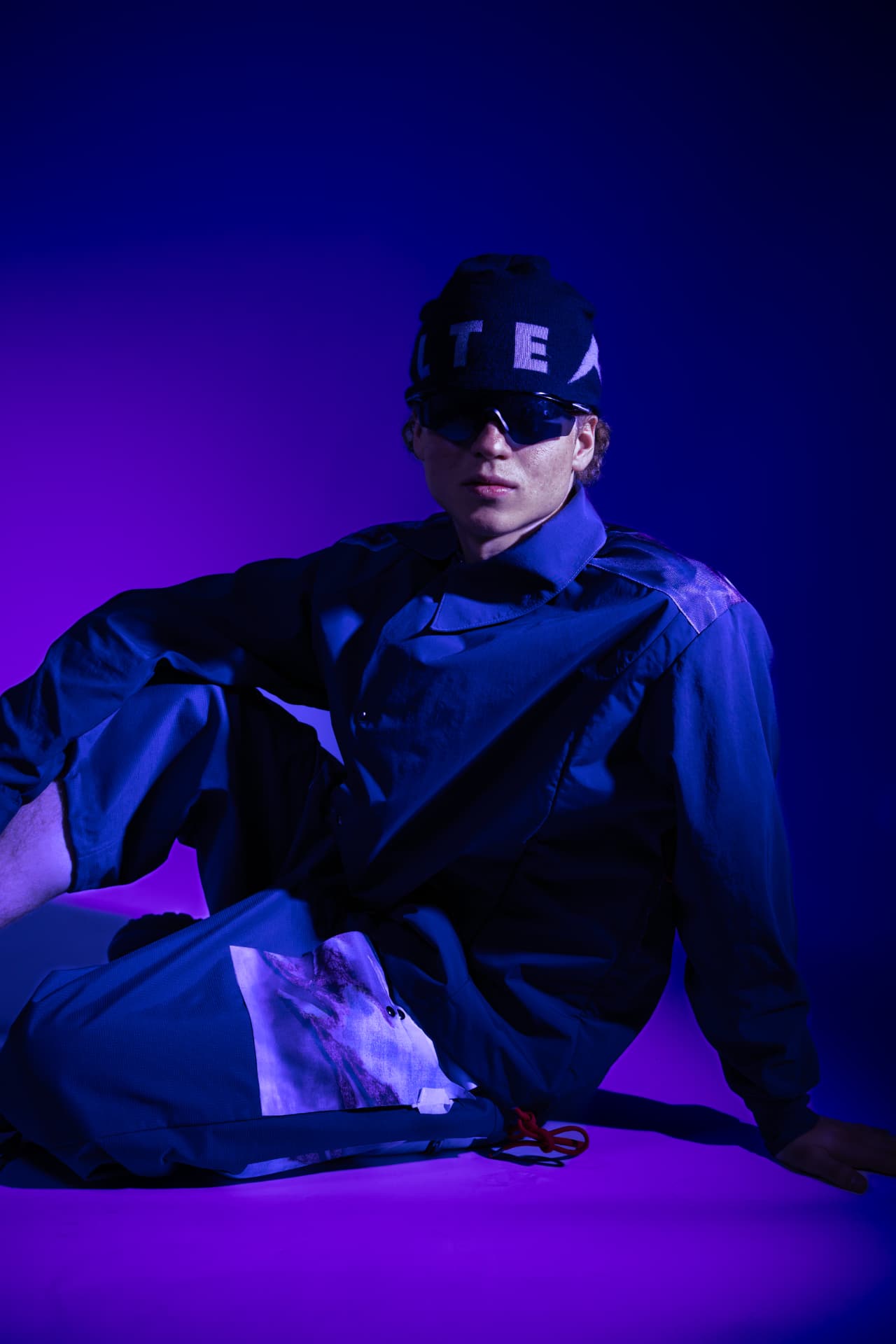 Model seated in a dark shell suit under purple light