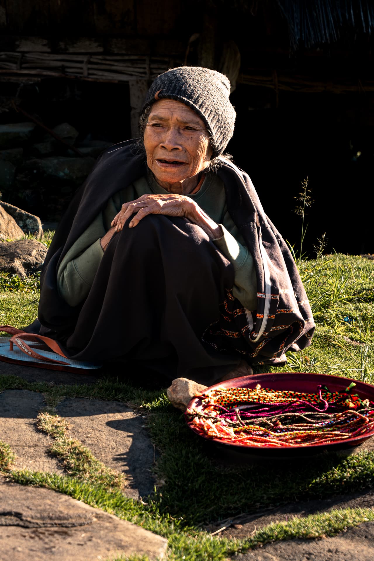 Elderly woman with a knitted hat seated beside a tray of beaded jewellery