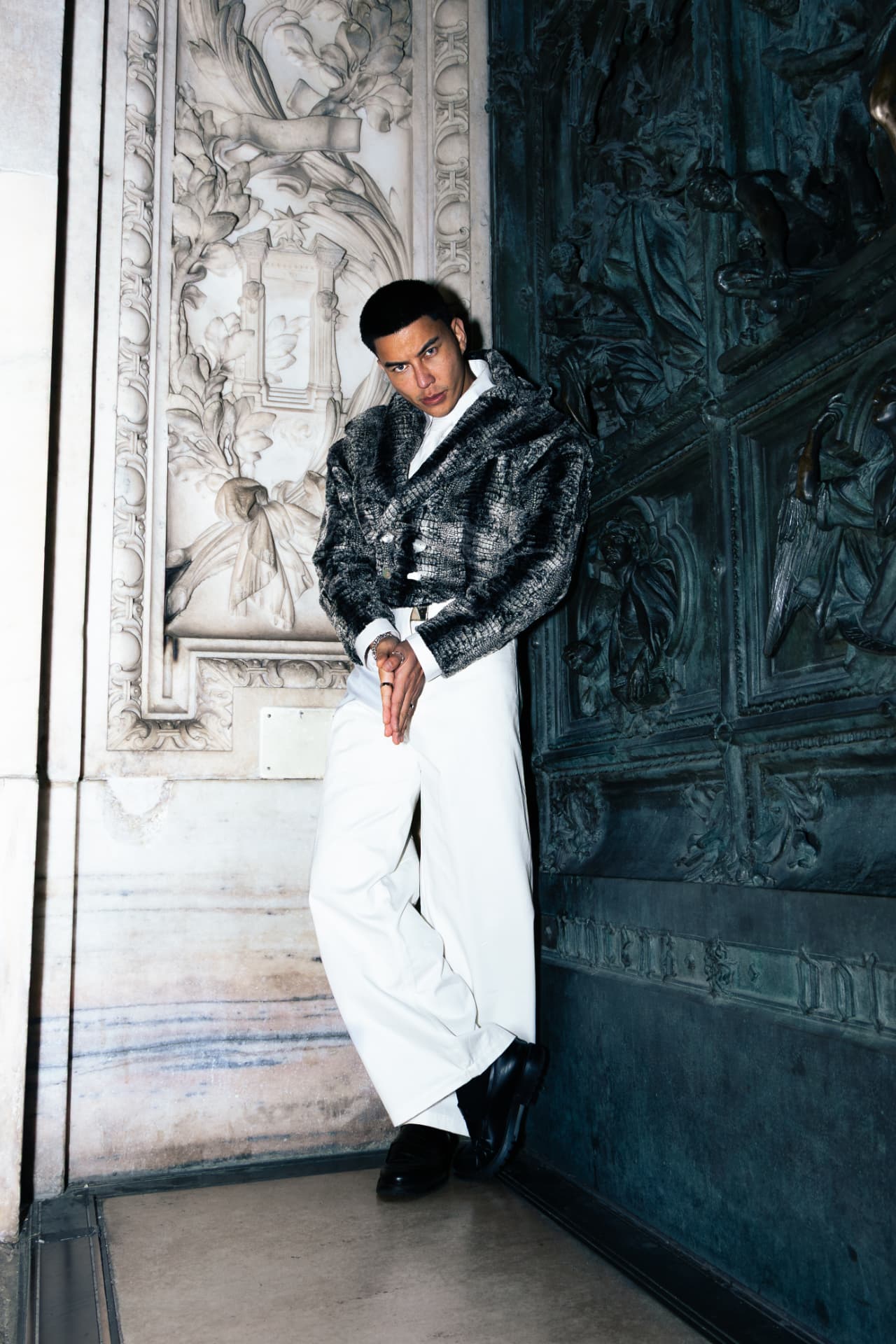 Model in a grey fur jacket and white trousers by bronze doors