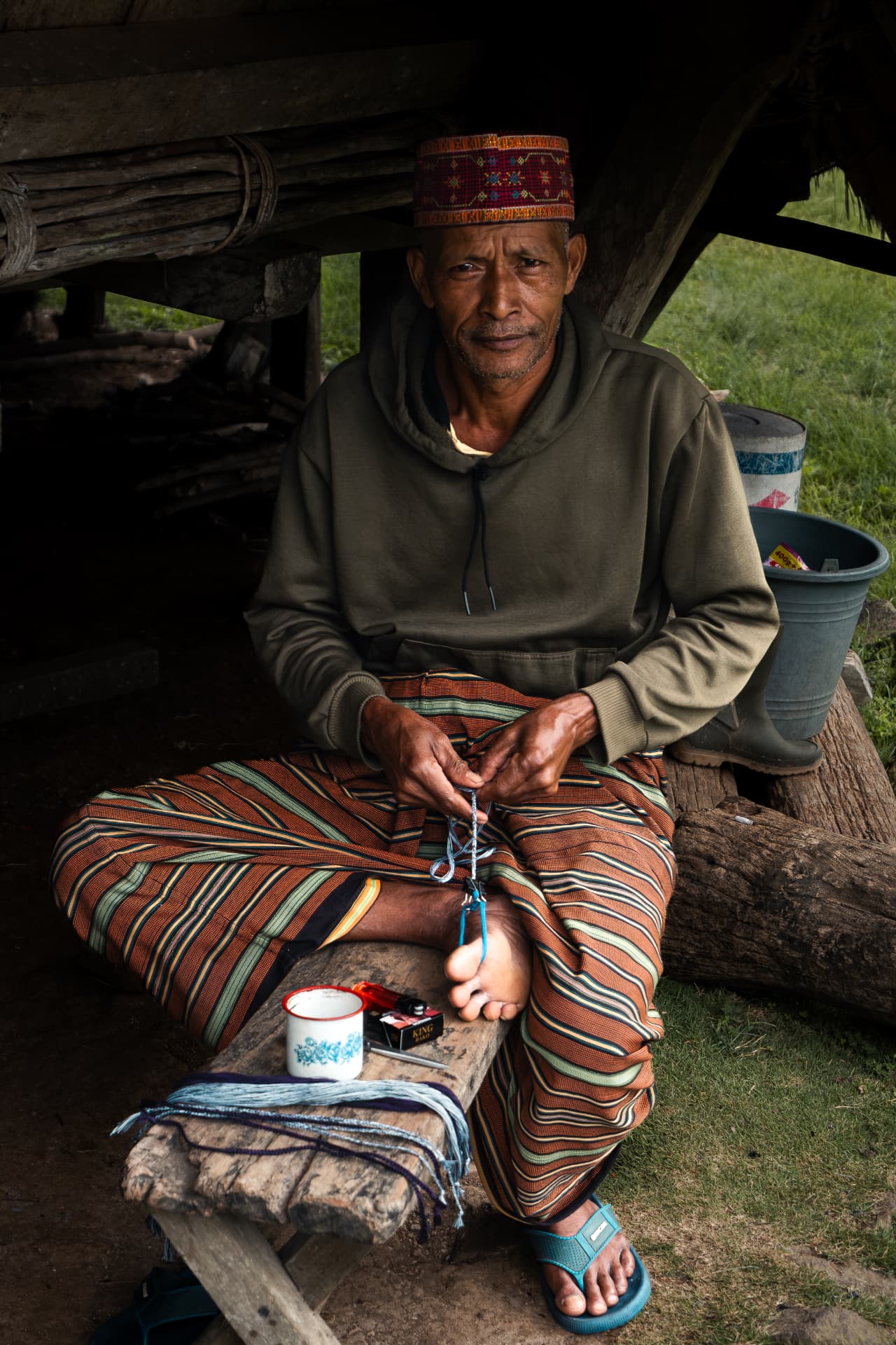 Man in a patterned hat weaving with blue string outside a wooden shelter
