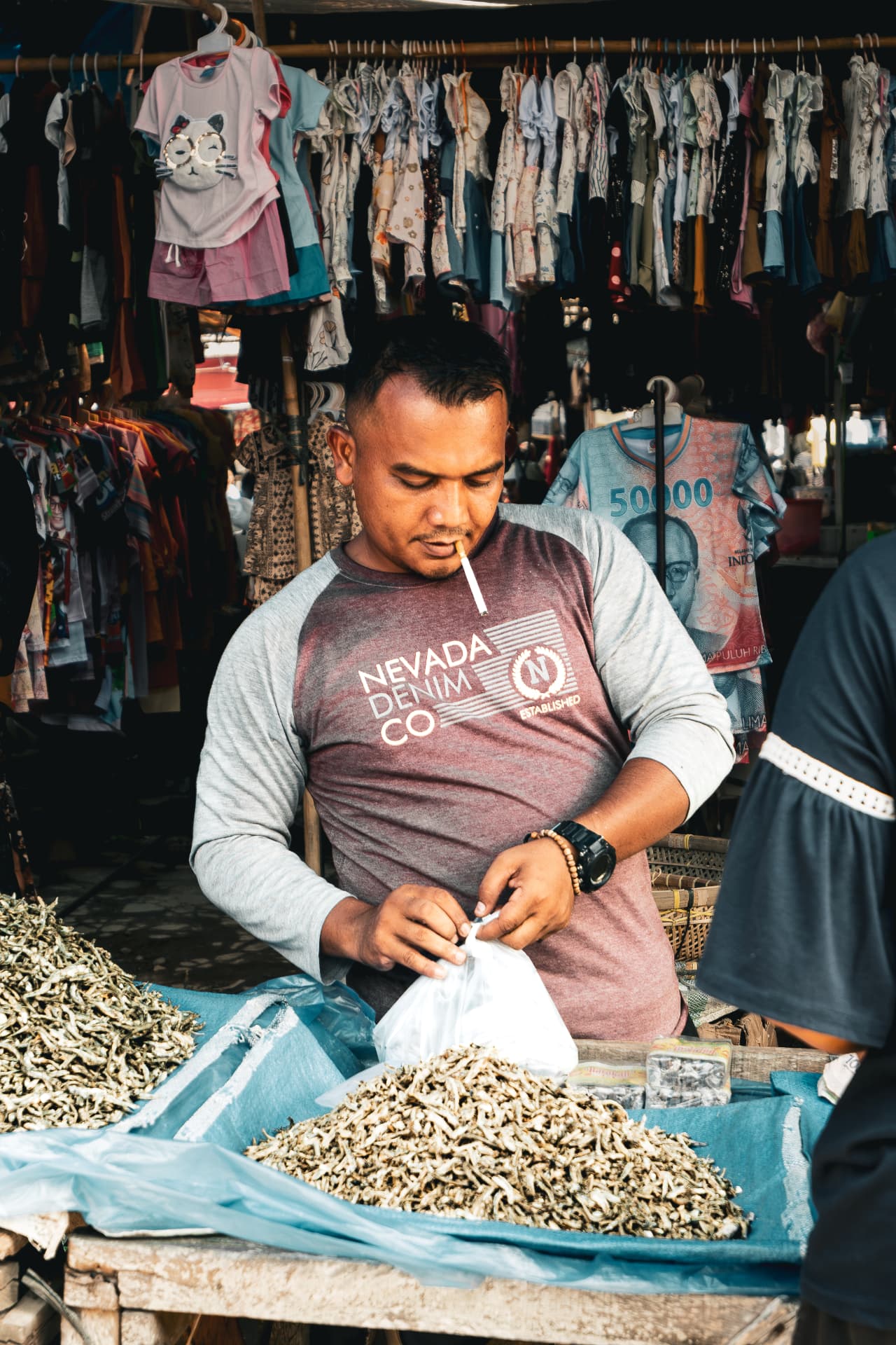Market vendor sorting dried seafood at a stall hung with children's clothes