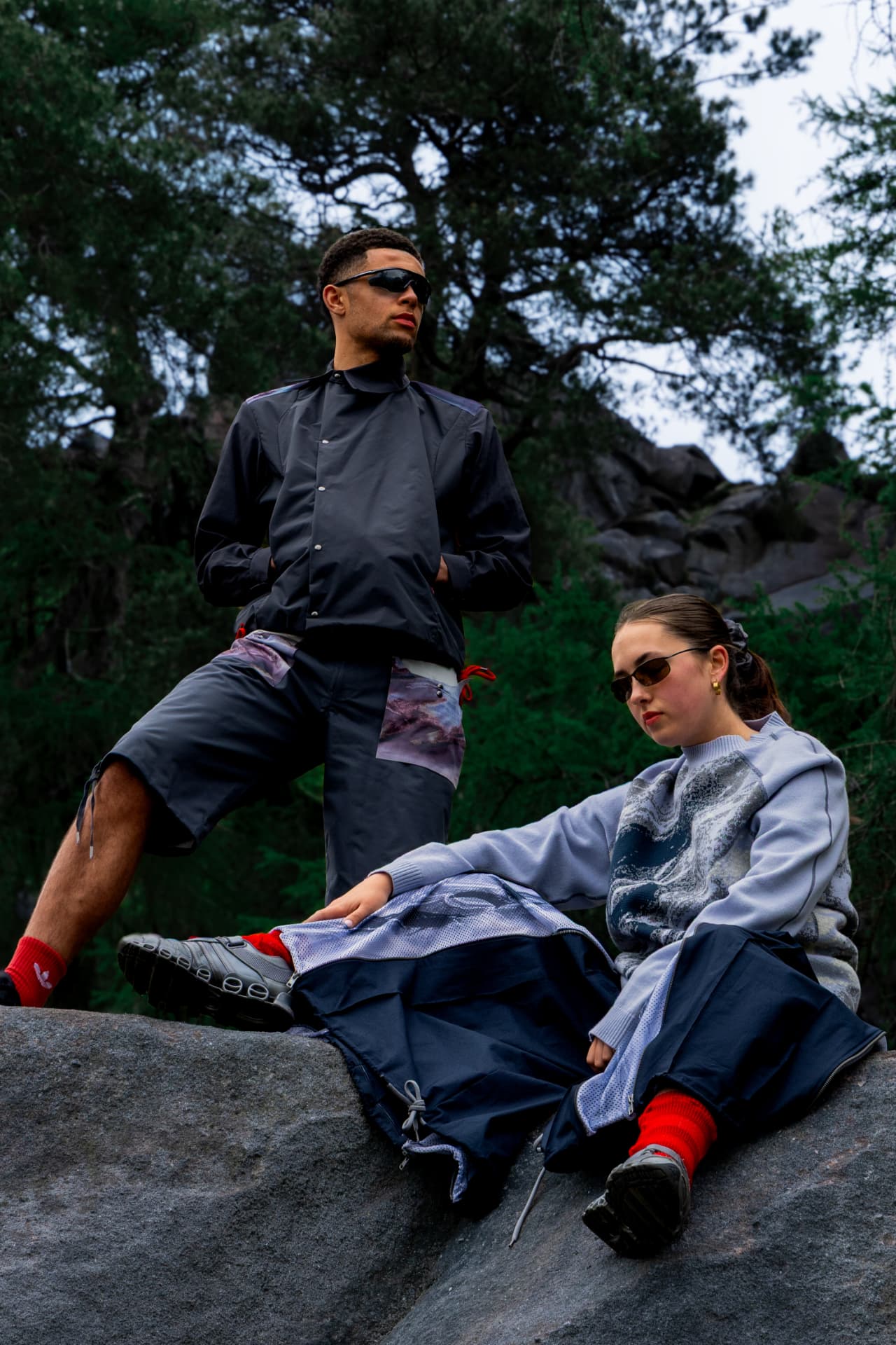 Two models on a boulder among pine trees in technical outerwear