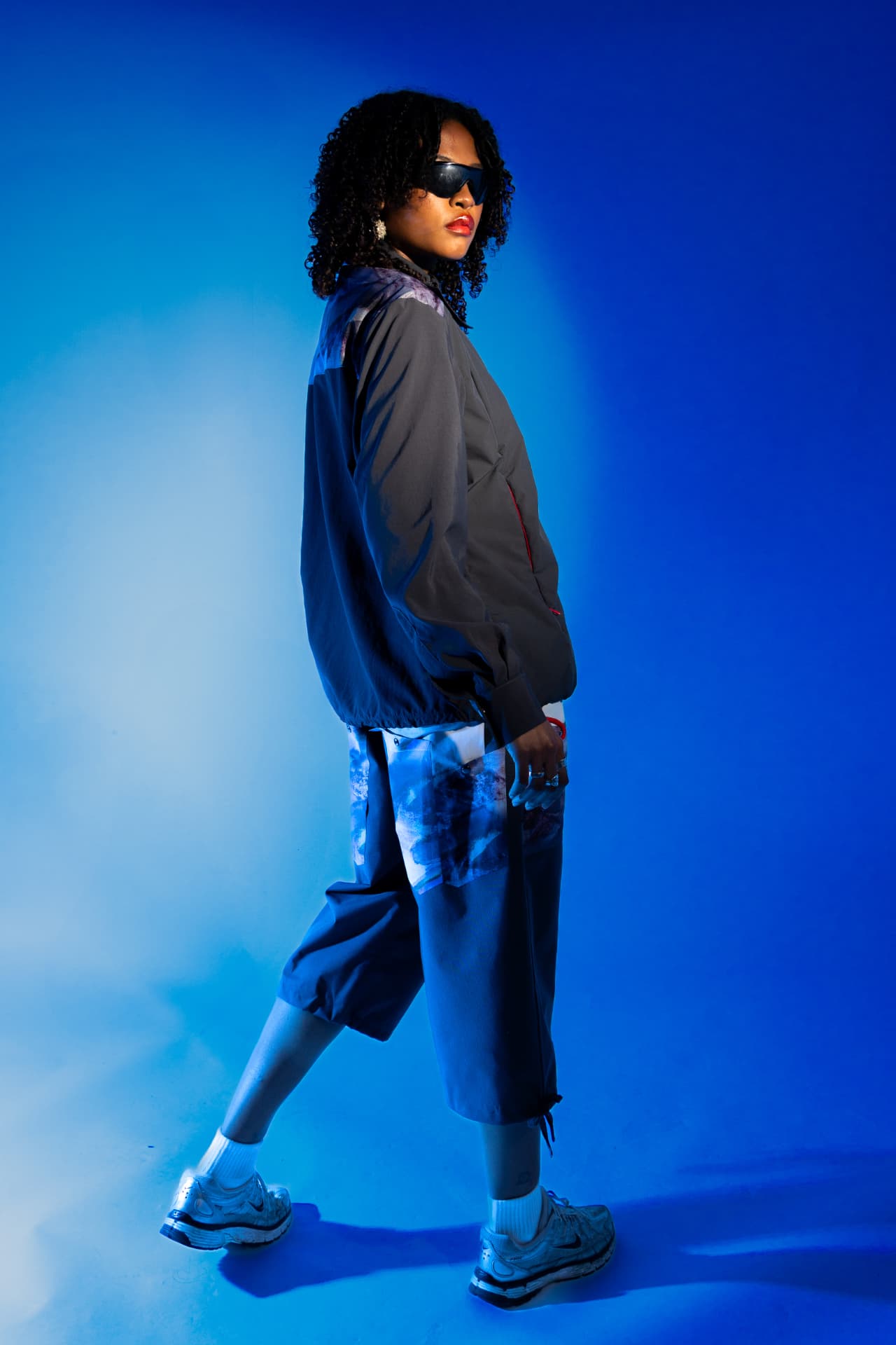 Model walking in a grey shell jacket against a blue backdrop